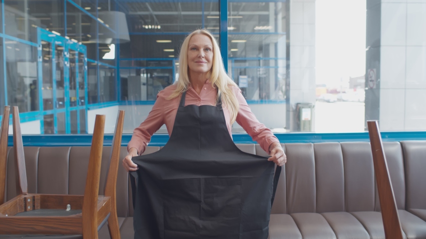 Senior happy cafe owner woman tie apron preparing to welcome guests in new cafe. Mature waitress smiling at camera excited about reopening of coffee shop after quarantine restrictions Royalty-Free Stock Footage #1057246702