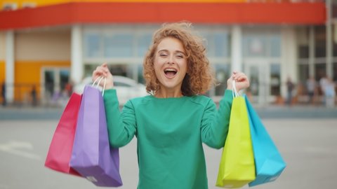 Portrait of cheerful young shopaholic woman holding paper bags with purchases and smiling at camera standing on parking lot of shopping center. Happy lady shopping on black friday