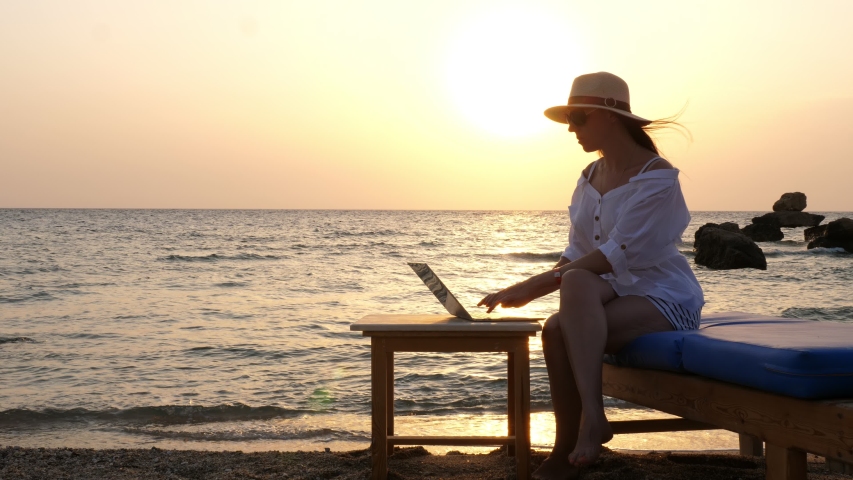 young woman in sunglasses and sun hat, uses laptop, sitting on the beach by the sea, at sunset or sunrise. freelancer, remote work. work and vacation at sea Royalty-Free Stock Footage #1057250668