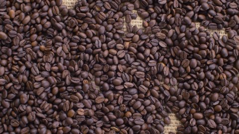 Close up roasted coffee beans on burlap. Hands placed down a a cup of coffee with froth. Camera slowly moving on slider, dolly shot, top view.