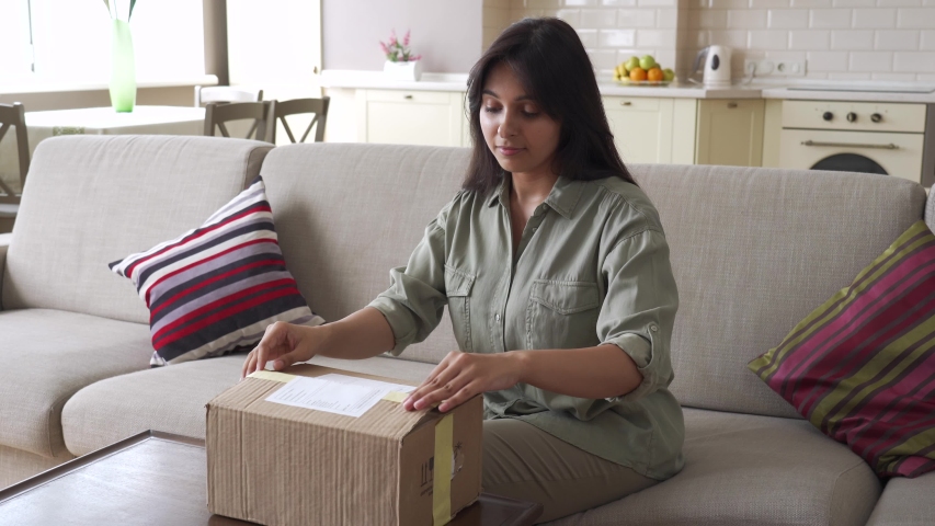 Happy indian young woman buyer opening parcel box at home. Smiling female customer shopper receiving online shop purchase buying clothes unpacking delivery postal shipping package gift sit on couch. | Shutterstock HD Video #1057255051