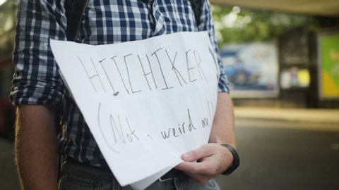 A hitchhiker holds a sign up asking for rides on the side of the street with cars and busses passing by in the blurred background.