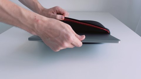 The guy putting small silver laptop into a bag and zipping it up on white background of the table and the wall. Action. Transporting a portable computer inside a protective folder.