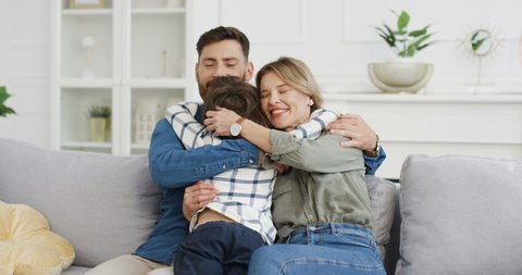 Rear of Caucasian cute little kid boy running to young happy mother and father sitting on couch in cozy living room. Cheerful parents hugging and embracing their small son at home on sofa. Back view.