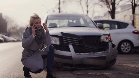 Young frustrated woman kneeling in front of the damaged car and calling for help after the car accident. Slow-motion shot with shallow focus and copy space