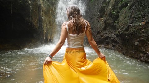 A young woman with long, dark hair is walking with her yellow pants / skirt into the water towards a small waterfall. Swinging and dancing with her body and arms to the sides.