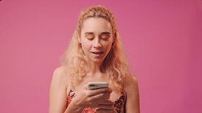Happy curly blonde young female holds mobile phone and makes okay gesture, expresses her approval, wears colored dress and bright makeup on pink background. Emotions and pleasant feelings concept.