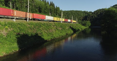 Ural Mountains, Bashkortostan / Russia - June 17 2020: Freight train carries with multicolored carriages an electric locomotive by two-sided Siberian railways near river in the forest mountains