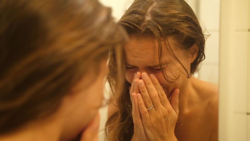 A woman with bruises and scars in her face from being punched and hit looks herself in the mirror in the bathroom and wipes the tears off her face. She breaks down and cries again.