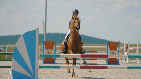 Competitive woman rider on horse jumping over obstacles, slow motion. Light-brown horse leaping fence on sandy parkour riding arena, equestrian competition outdoors. Training jumping hurdle
