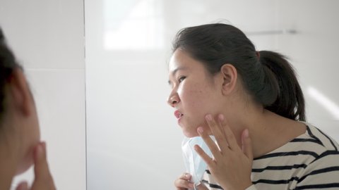 Young adult asian woman worry unhappy with skin acne or pimples from wearing face mask showing swollen, spot, scar, skin allergy on chin and cheek in mirror in maskne covid-19 social distance concept.