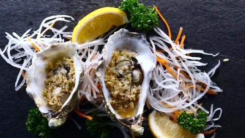 Waiter Is Serving Fresh Raw Oysters in Half Shells with Lemon and Fried Garlic on Black Textured Slate Background. Top View, Close-up. Woman Hand Puts Oyster on the Table. Seafood Dinner in Restaurant