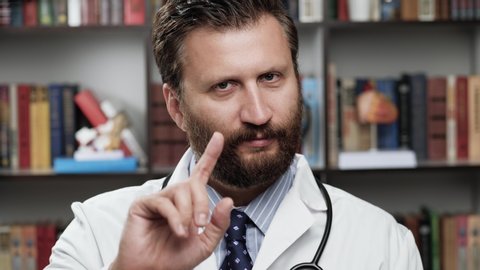 Doctor says no prohibitively points his finger. Strict male doctor in white coat and stethoscope in office looks at camera, raises his hand and begins to show disapprovingly with his index finger