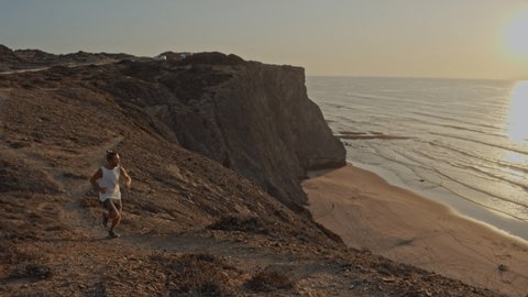 Slow motion shot of man running on cliff above beach. Young man jogging in nature at sunset, ocean views from above 