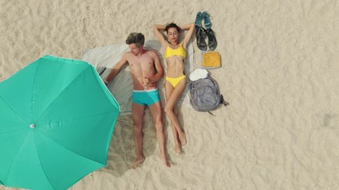 relaxed couple lying on white sand under turquoise sun umbrella on empty wild beach at seaside on summer vacation.  Aerial drone footage