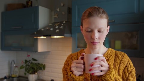 Portrait young woman drinking tea in kitchen. Attractive woman drinks warming Cup coffee at home in cool weather. Smiling woman in sweater holding cup tea in hands at morning and look at camera วิดีโอสต็อก