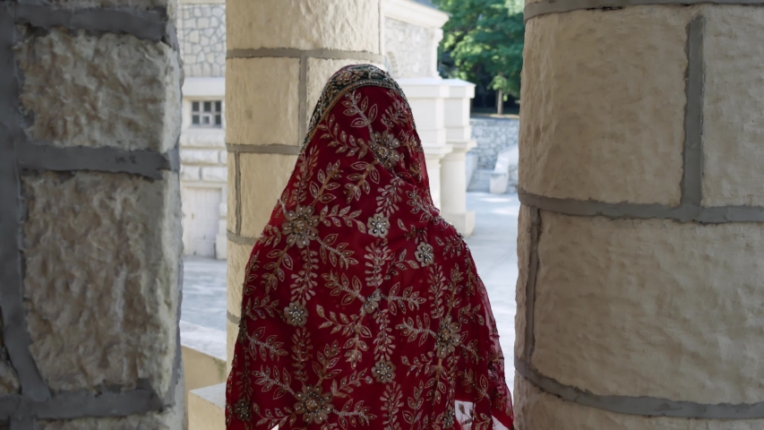 From behind Indian woman walks on streets of ancient architecture city of India dressed in red Sari, decorated with traditional ornaments and Mehendi patterns henna drawings on hands.
