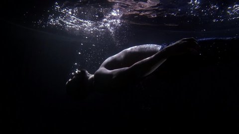 Close-up of a bearded man falling back into the dark water, he is in his pants and with a naked torso. There is darkness and bubbles around it, and a little light comes from the surface of the water