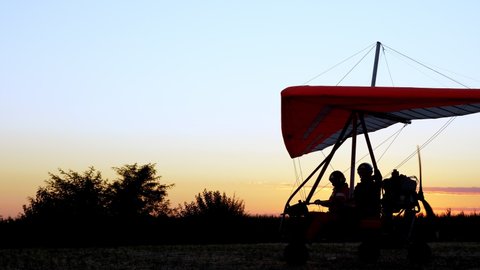 at sunset, dark silhouette of motorized hang glider with passenger and pilot preparing for takeoff