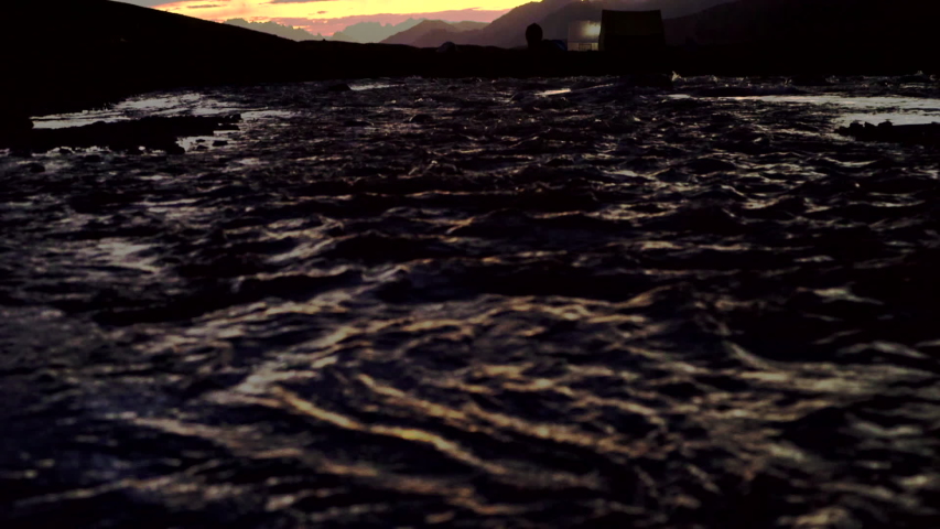 Tilt up dark shot from over a river to an amazing sunset in the mountains with silhouettes of tents, orange and blue colours painted the sky.