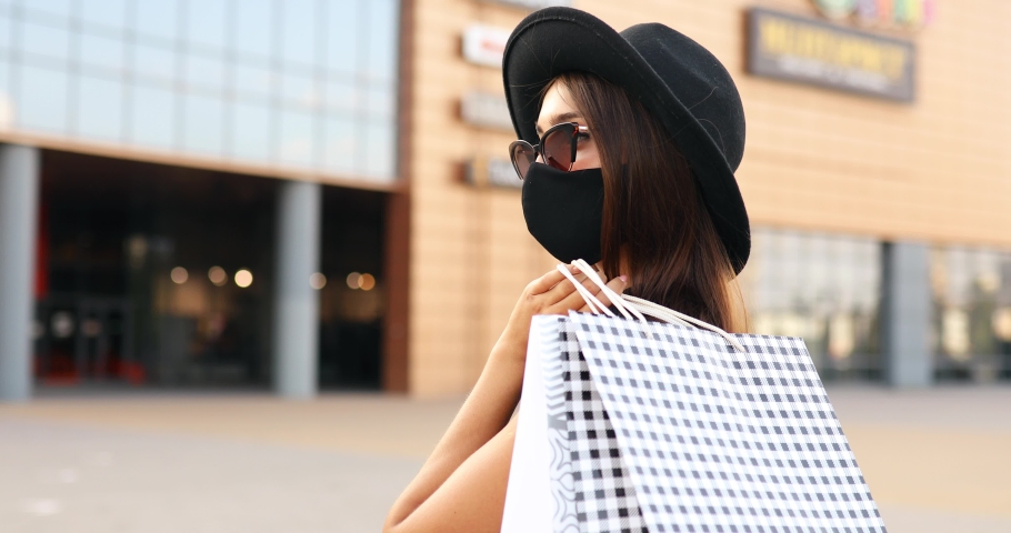 elegant stylish Shopaholic girl in a medical protective mask on her face, in a black dress and hat, shopping in a shopping center, with shopping bags. Black Friday, shopping Royalty-Free Stock Footage #1057306435