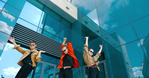 Group of three young crazy dancing people actively moving together, waving hands, modern choreography on city street. Two boys and one teen girl jumping funny moving together, urban bright dance