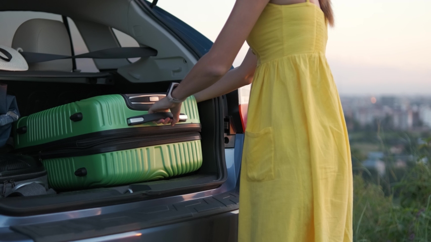 Young woman in summer dress taking green suitcase from car trunk. Travel and vacations concept. | Shutterstock HD Video #1057308463