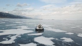Aerial view of a vehicle with rubber air cushion on Siberian lake Baikal in winter. Clip. Rear view of a khivus boat sliding fast on ice and snow on forested mountains and blue cloudy sky background.