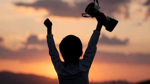Celebrating victory winner win success concept. Silhouette of happy sport little champion sportsman, sporty little boy child holding up golden trophy cup in hand against sky raised hands. 4 K slow-mo