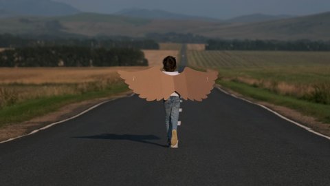 Happy little boy child angel running with toy cardboard plane wings on rural road. Kid aviator pilot of airplane dreams of flying, 4 K slow-mo