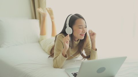 Young Asian woman prone on the bed listening to music through her headphones and laptop.
