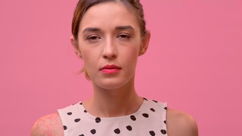 Close up portrait of an angry caucasian female with black tied hair shouting WTF and grabbing her hands behind her head on a pink background. Blaming, warning, accusing concept.