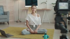 Wide shot of young Caucasian sportswoman wearing grey leggings and white t-shirt is sitting on mat in front of camera and telling about training purpose
