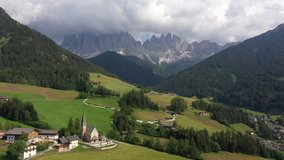 Retreating aerial footage flying over church of small village (Santa Magdalena) in the Italian Alps (Dolomites), religious landmark and natural landscape in Europe