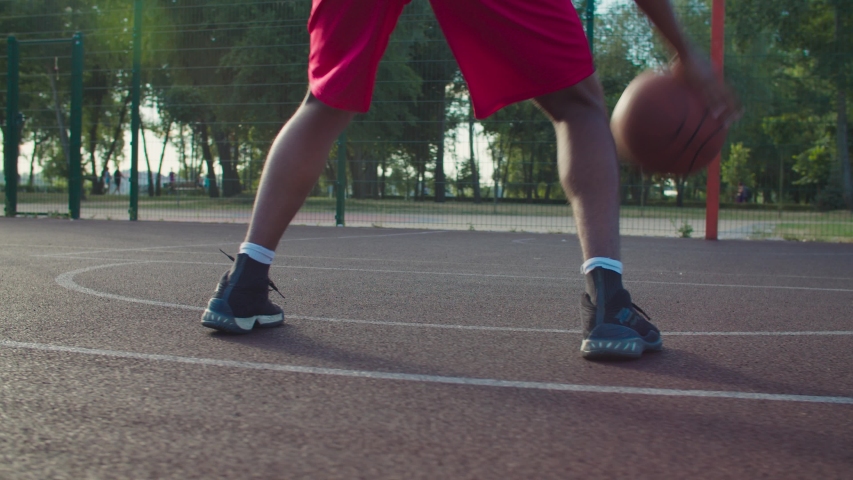 Rear view of active male basketball player practicing ball handling skill, dribbling ball between legs on outdoor court in rays of early morning rising sun during streetball training in neighborhood. Royalty-Free Stock Footage #1057324438