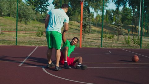 Sporty fit mixed race streetball player extending hand to lift fallen opponent player off ground, helping to stand up , practicing good sportsmanship during basketball game on outdoor court.