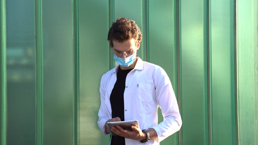 Caucasian young male doctor wearing face mask while using digital tablet against green wall during COVID-19 outbreak. High quality 4k footage