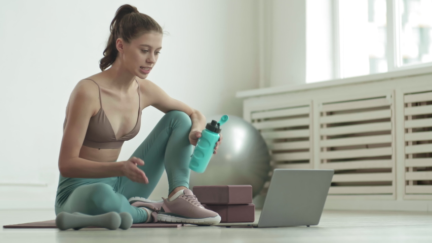 Lockdown of young Caucasian trainer wearing stylish sportswear sitting on mat in front of laptop, drinking water from sport bottle and speaking