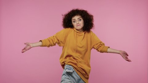 Poor student girl afro hairstyle in hoodie showing empty pockets in jeans pants, doing no money gesture, debt problems, financial crisis and bankruptcy. indoor studio shot isolated on pink background