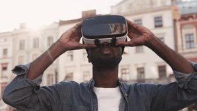 Young African American man playing in virtual reality headset in city center. Guy is using VR glasses and smiling.