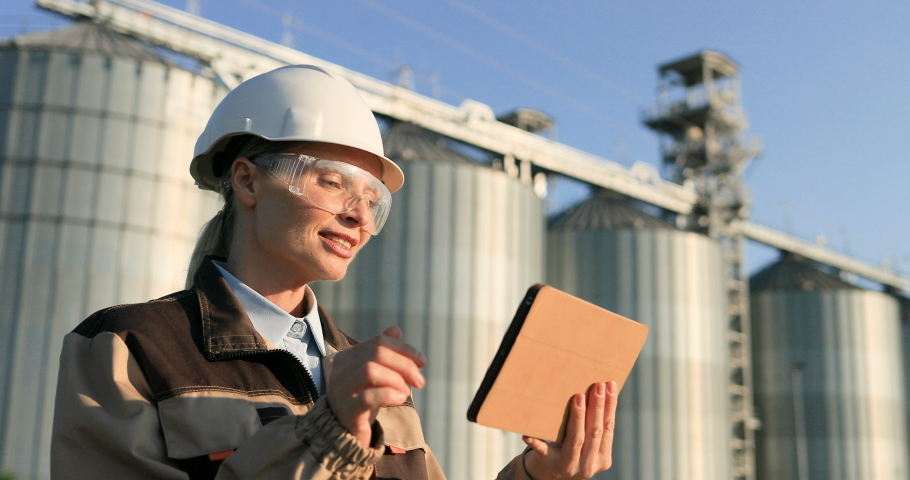 Side view of beautiful happy female builder having video chat on tablet at work. Close up portrait of woman worker in helmet and goggles videochatting on device outdoors in good mood. Factory concept