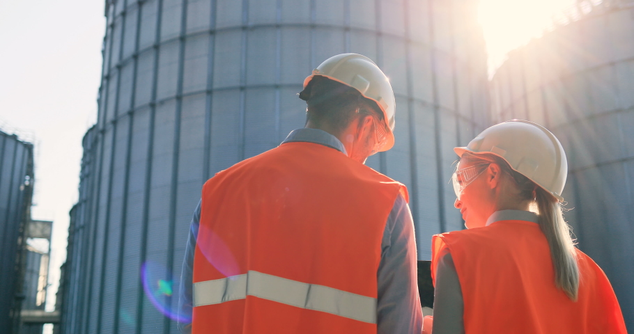 Rear of male and female Caucasian engineers in helmets and uniform standing outdoors at plant near big cisterns and talking. Woman and man controllers looking at away and discuss industry project