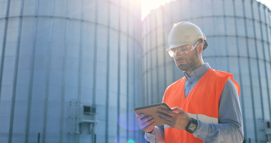 Close up portrait of joyful Caucasian male engineer in uniform and helmet standing at plant outdoors with tablet. Man in goggles standing near big cisterns and smiling to camera. Worker concept