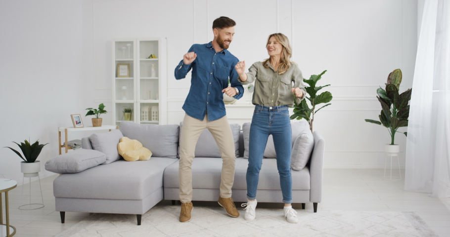 Joyful Caucasian young funny married couple having fun and dancing in cozy living room. Beautiful happy woman and handsome man dance and laughing at home. Wife and husband relationships. Indoors. | Shutterstock HD Video #1057332613