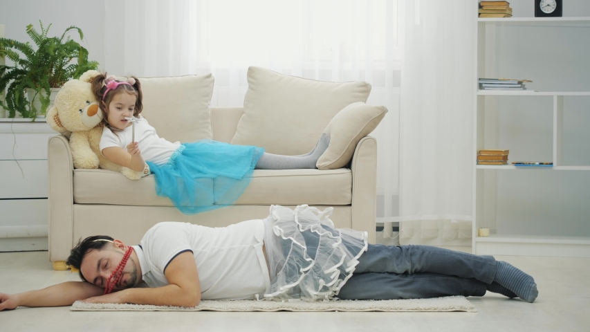 Cute girl is waving magic wand, and daddy in funny clothes is obying her commands, lying on the floor, completely tired. | Shutterstock HD Video #1057334269