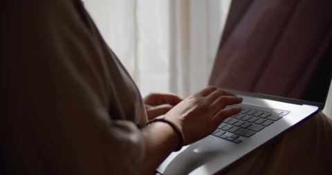 Beautiful warm sunlight peeks through window and sheer curtains as woman writes an email quickly on her laptop. Close up of hands typing on computer while working from home