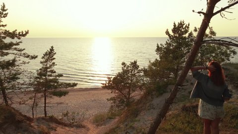 Drone flight from backside to sea horizon. Young woman enjoys beautiful scenic sunset at Baltic Sea standing at tree on hill
