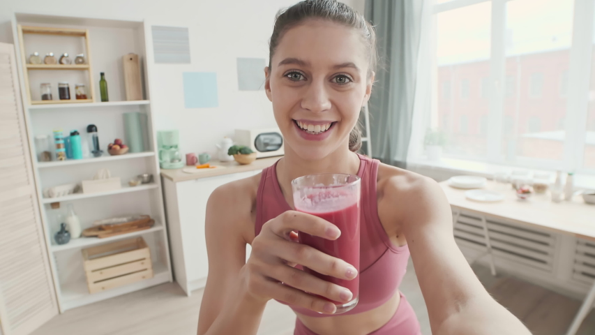 Medium shot of young Caucasian sportswoman standing in the kitchen, holding camera in her hand and glass with fresh yogurt in another, making sip and telling about its benefit | Shutterstock HD Video #1057342990