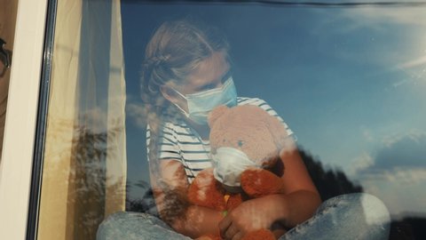 bored kid in medical mask in home quarantine coronavirus sitting by the window. child with a toy teddy bear in protective masks looking out the window. coronavirus epidemic covid 19 prevention concept
