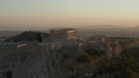 Aerial Flight towards Acropolis of Athens with Greek Flag Waving in Beautiful Golden Hour Sunset light with Ocean in the distance